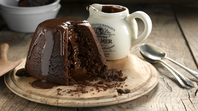 Steamed Chocolate Pudding covered in chocolate sauce, served on a board next to a white jug of chocolate sauce