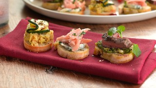 Three party crostinis served on a napkin in front of a platter of more crostinis.