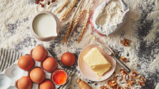 Baking scene setter with wheat, a jug of milk, open flour bag, 6 eggs with one cracked open and butter.