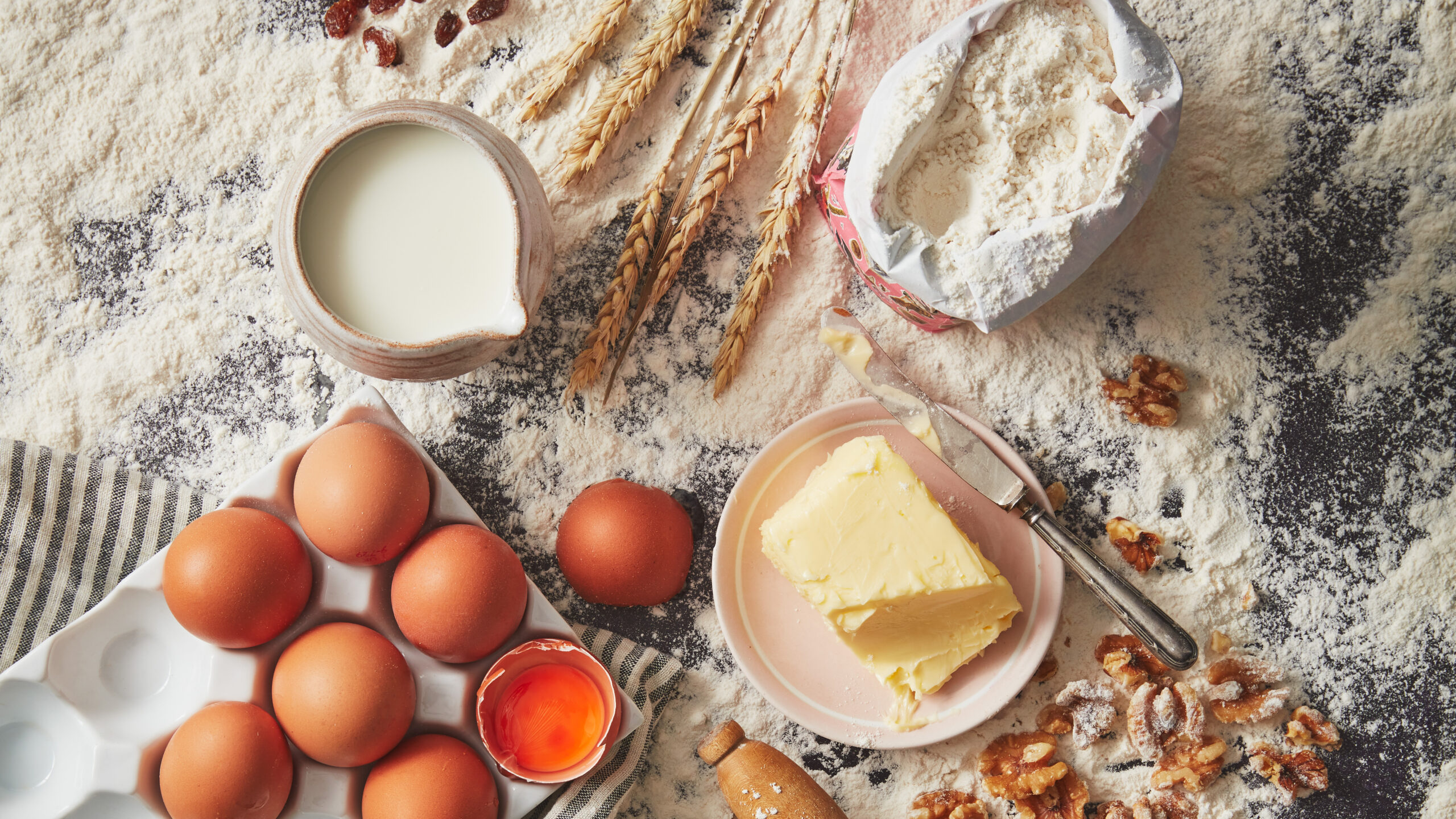 Baking scene setter with wheat, a jug of milk, open flour bag, 6 eggs with one cracked open and butter.