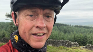 A close up of Darren's face, with the countryside in the background. Darren is wearing a cycling helmet and smiling.