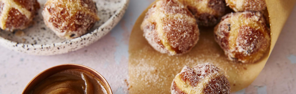 Hot Cross Bun Doughnuts wrapped in a brown paper cone, in a bowl, with a small pan of caramel sauce