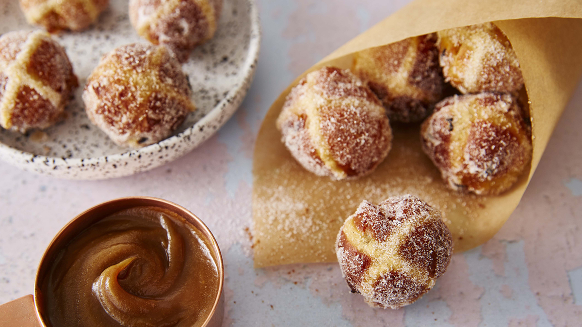Hot Cross Bun Doughnuts wrapped in a brown paper cone, in a bowl, with a small pan of caramel sauce