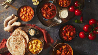 An overhead view of 6 curries laid out on a dark background surrounded by naan breads and key ingredients such as tomatoes, ginger and chillies