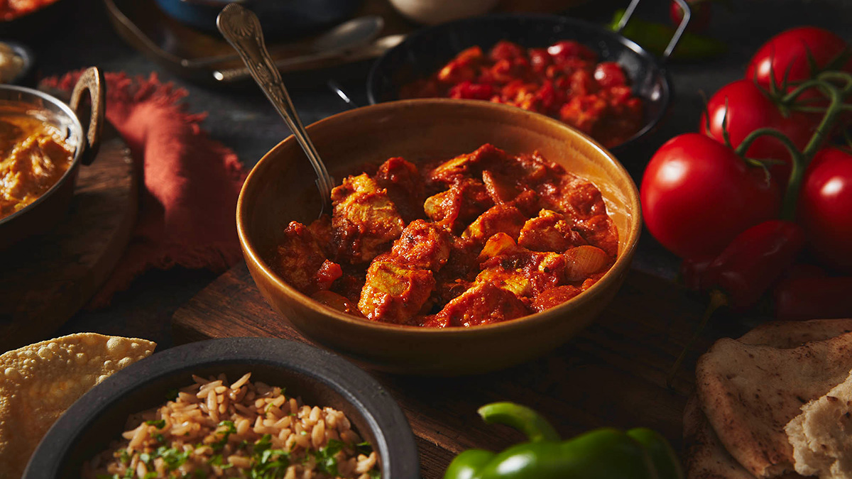 A close up of curry in a bowl with a spoon, to the back is another blurred out curry, with tomatoes to the right and a bowl of rice at the bottom.