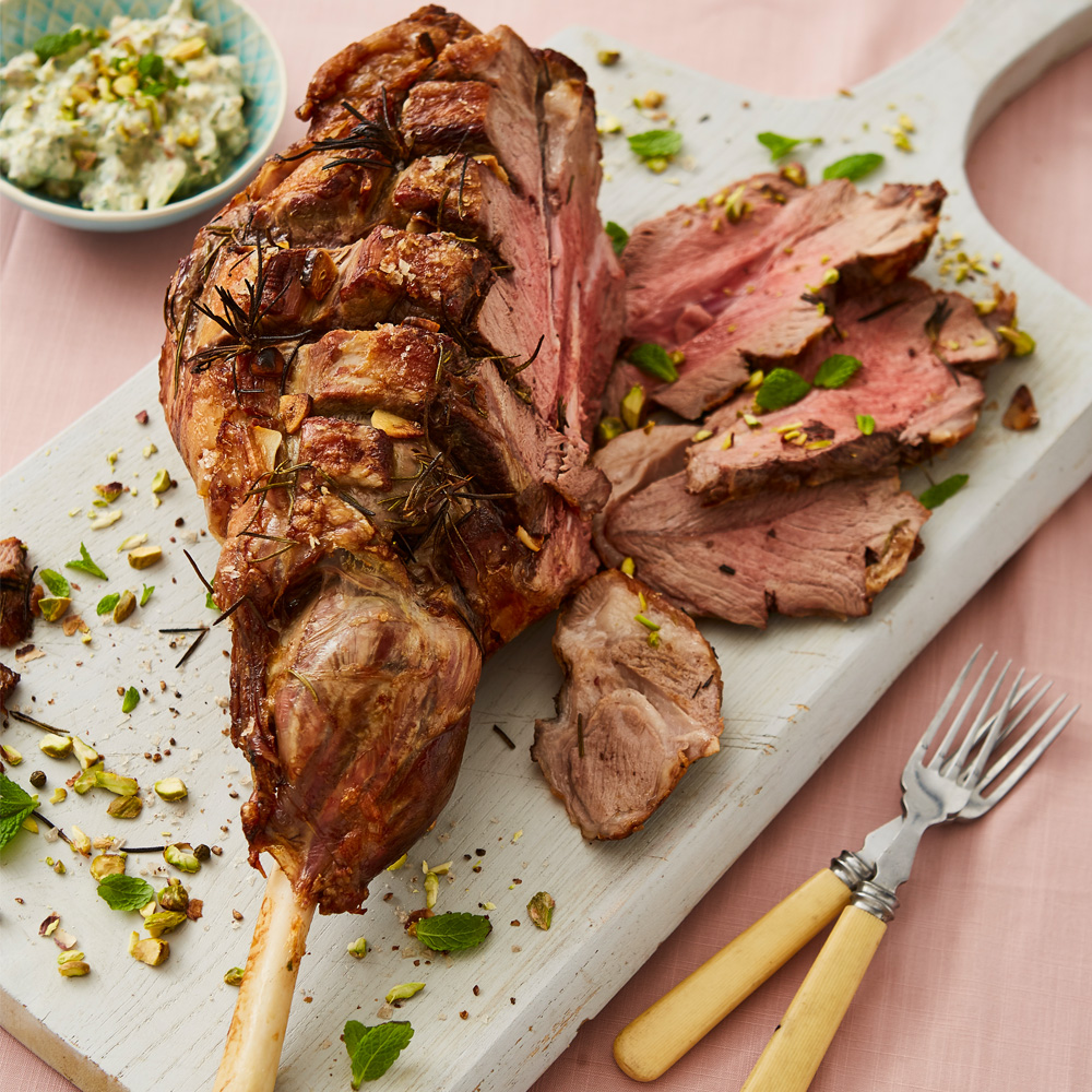 Image of a roast leg of lab with herbs on top on a chopping board with a pink background