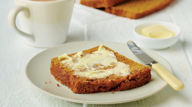 A slice of Granny Hilton's Sticky Bread on a white plate, with butter spread on top and next to a cup of tea