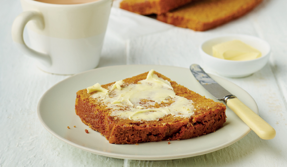 A slice of Granny Hilton's Sticky Bread on a white plate, with butter spread on top and next to a cup of tea