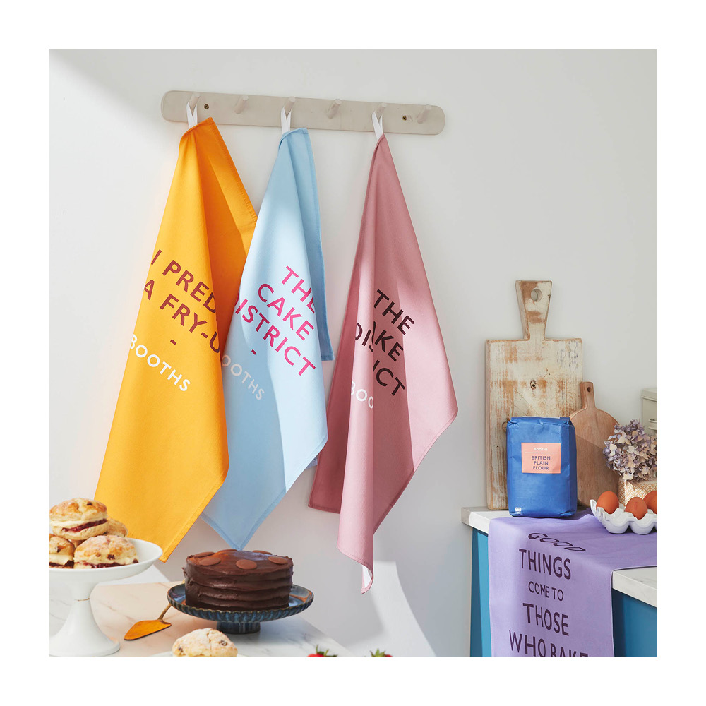 Image of 3 Booths Pun tea Towels hung up on pegs in a kitchen. One is yellow, one is blue and one is pink.