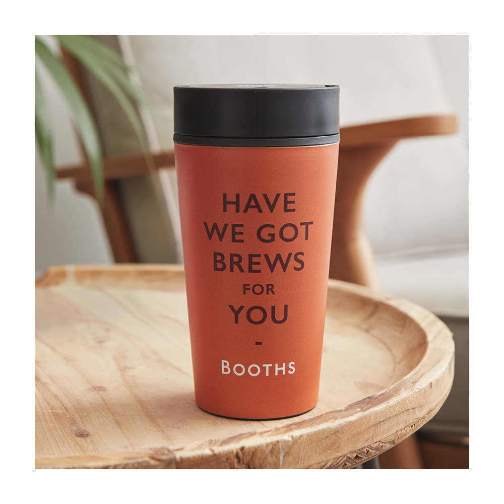 An image of a Booths Pun Travel Mug sat on a table. It's orange and says 'have I Got Brews For You' on the front