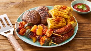 Plate of BBQ food including grilled corn, sausages, burgers and kebabs