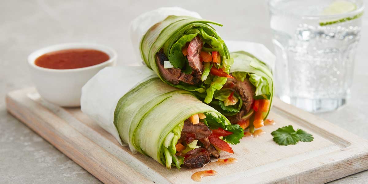 Three Roast Beef Rolls stacked on top of one another on a wooden board, next to a bowl of sweet chilli sauce