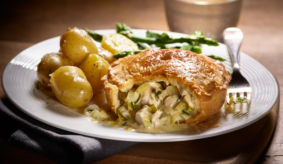 Chicken, leek and tarragon pie, served on a white plate with new potatoes and asparagus, with a slice missing to show the filling