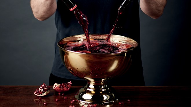 Sangria in a golden serving bowl, with red wine being poured in and pomegranates to the side