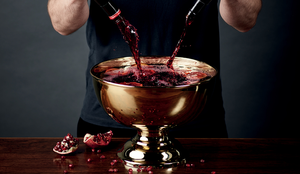 Sangria in a golden serving bowl, with red wine being poured in and pomegranates to the side