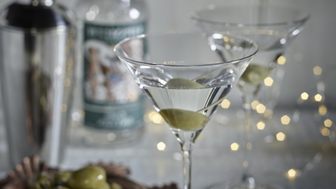 Two Classic Martinis served in glasses with an olive and white fairy lights in the background
