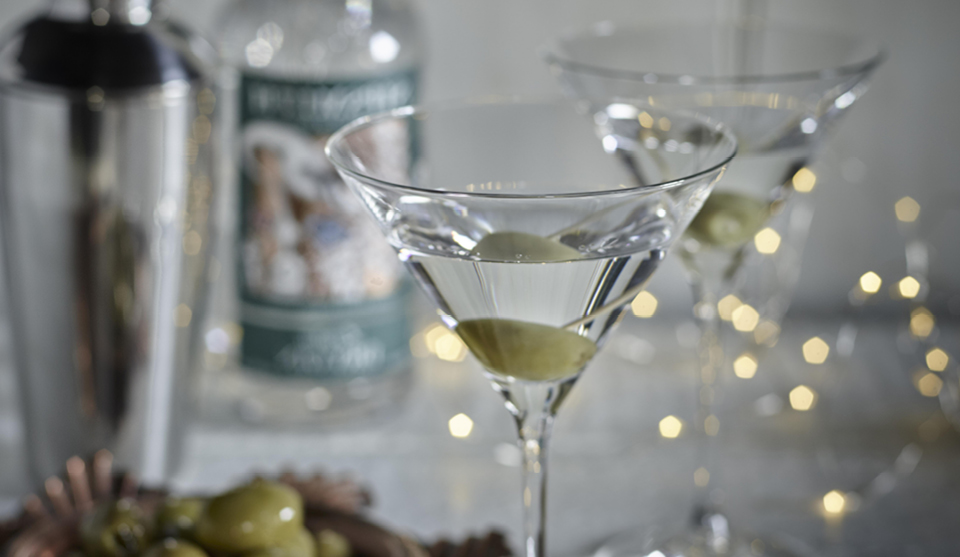 Two Classic Martinis served in glasses with an olive and white fairy lights in the background