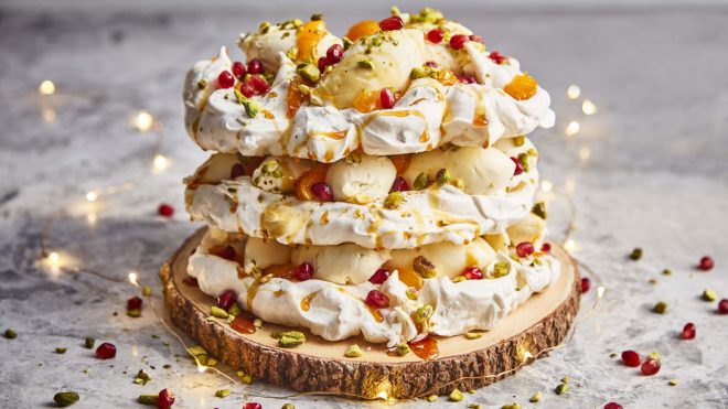 Pomegranate and Clementine Pavlova presented on a wooden board surrounded by pistachios and pomegranate seeds