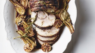 Boxing Day Porchetta served on a white dish, surrounded by roasted fennel and garlic bulbs. The porchetta has been sliced