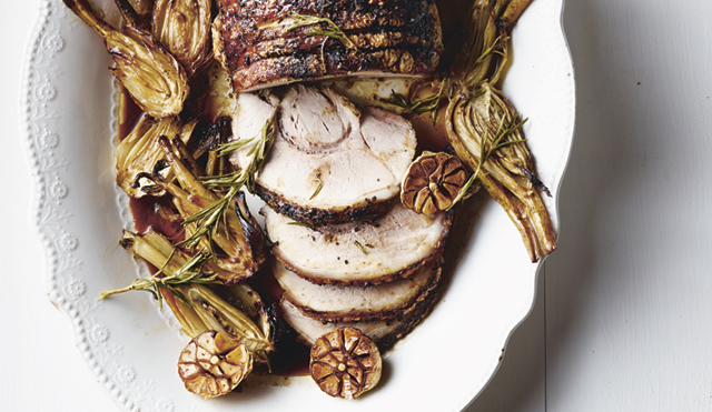 Boxing Day Porchetta served on a white dish, surrounded by roasted fennel and garlic bulbs. The porchetta has been sliced