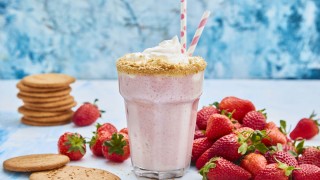 Strawberry Cheesecake Shake served in a glass with two straws, next to a pile of strawberries and digestive biscuits