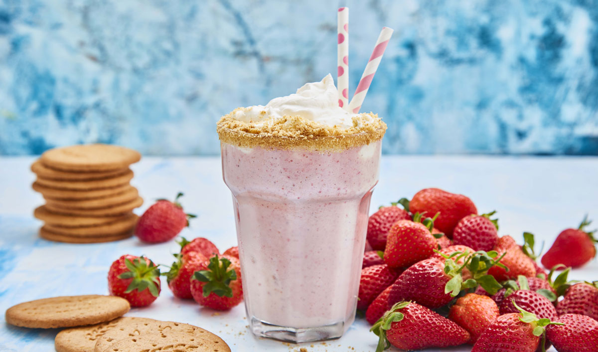 Strawberry Cheesecake Shake served in a glass with two straws, next to a pile of strawberries and digestive biscuits