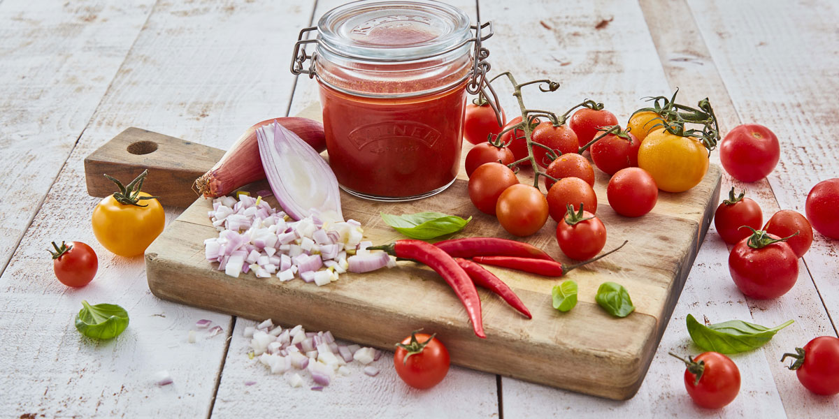 Tomato and Chilli Passata served ina glass jar, surrounded by vine tomatoes, chillies and onions.