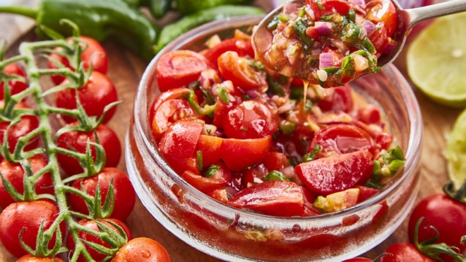 Tomato, Lime and Jalepeno Salsa served in a glass bowl, with a spoon lifting a portion out, surrounde by vine tomatoes and lime wedges.