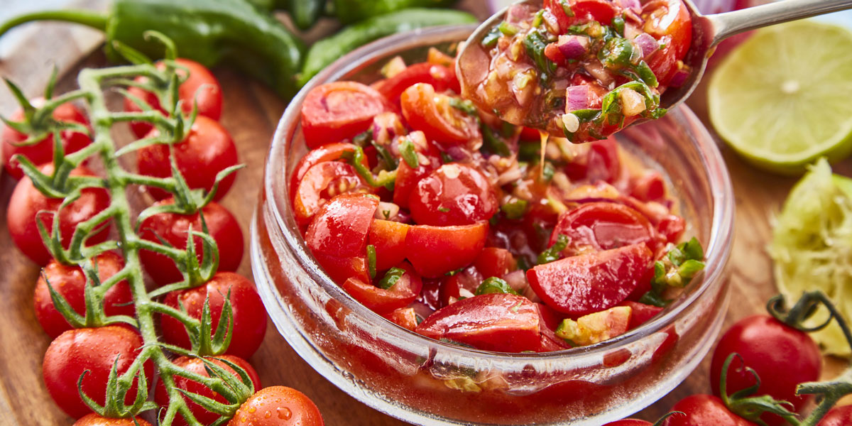 Tomato, Lime and Jalepeno Salsa served in a glass bowl, with a spoon lifting a portion out, surrounde by vine tomatoes and lime wedges.