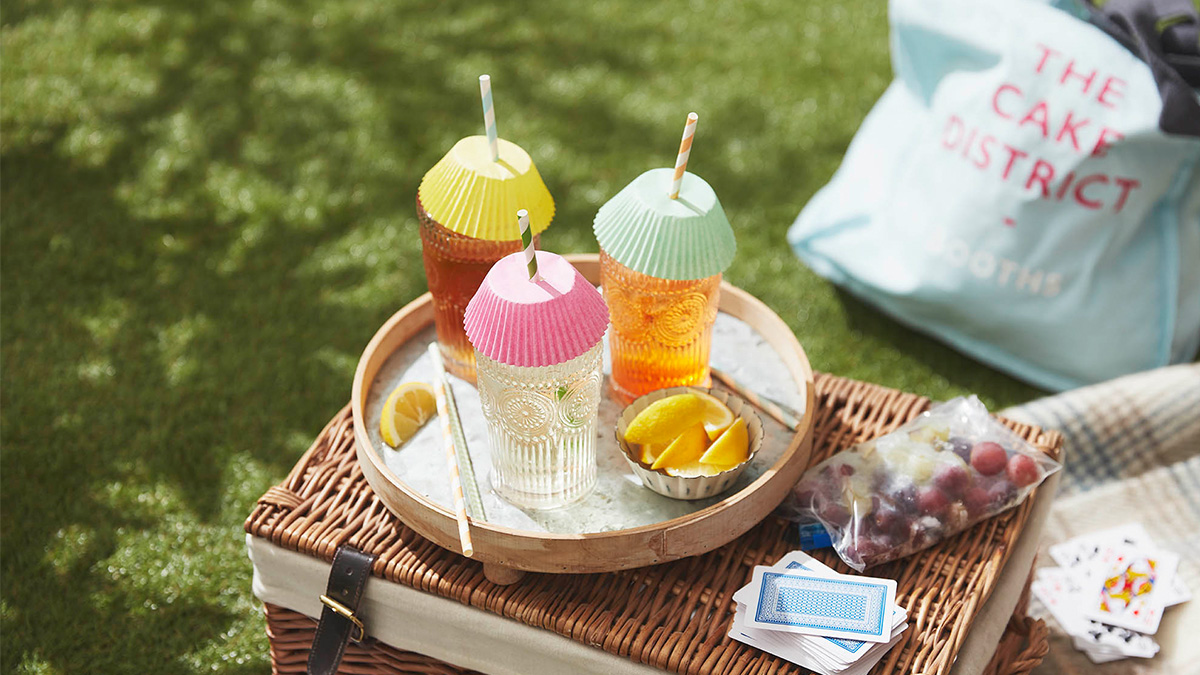 Image of drinks sat on top of a picnic hamper with a card game and Booths pun bag in the background.